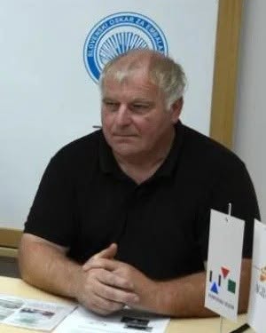 Moški z sivimi lasmi sedi za mizo, v ozadju je znak z modro in belo barvo.